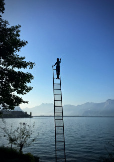 Mensch steht oben auf einer Leiter nahe dem Himmel, vor einer Bergkulisse mit See